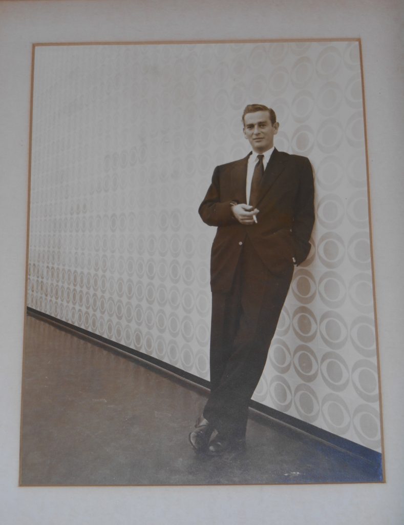 Portrait of man in a suit, leaning against a wall patterned with CBS logo. 
