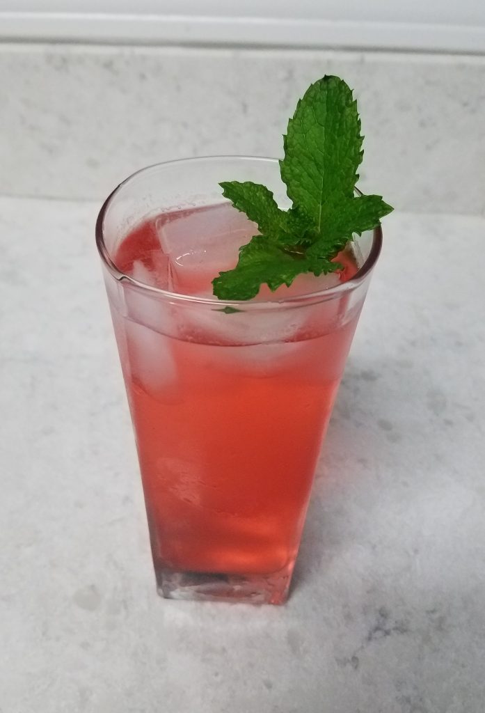 photograph of completed shrub drink in a glass with ice. The drink is pink, and there is a mint sprig as a garnish. 