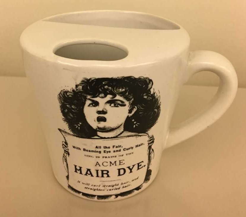 Photograph of a mustache cup. It is a mug with a semicircular ledge inside. The ledge has a half moon-shaped opening to allow the passage of liquids and serves as a guard to keep moustaches dry. The side of the mug features an advertisement for Acme Hair Dye. 