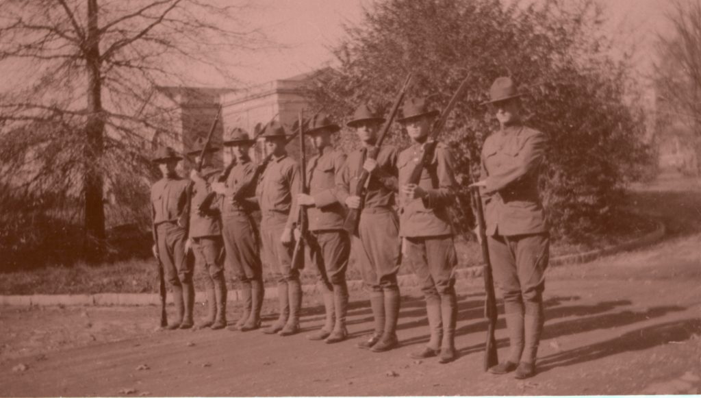 Black and white photograph of young men in Army uniform. They are standing in a line together, holding rifles. A building on Duke's East Campus is in the backgroun
