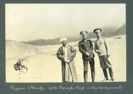 Three Russian officials standing together, a camel passes by in the background.