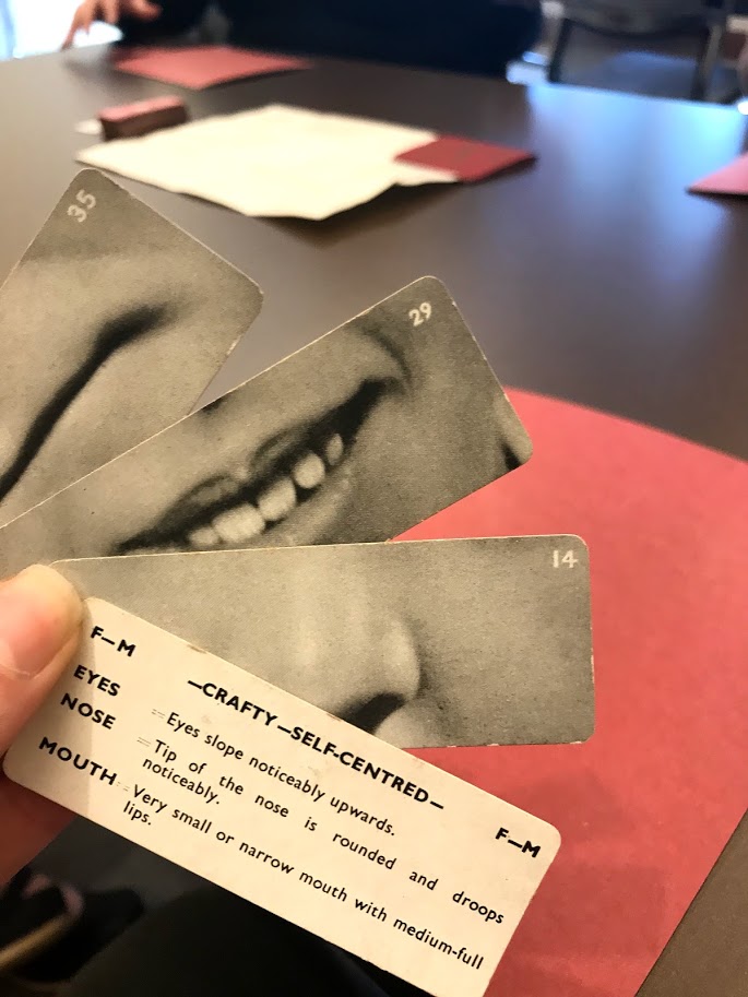 Photo of someone's hand holding four cards. One with a nose on it and two with a mouth, and a fourth with a description of a "crafty - self-centered" face.