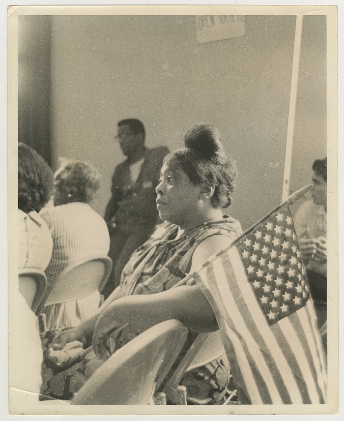A black and white photo showing Fannie Lou Hamer seated at an event holding an American flag.