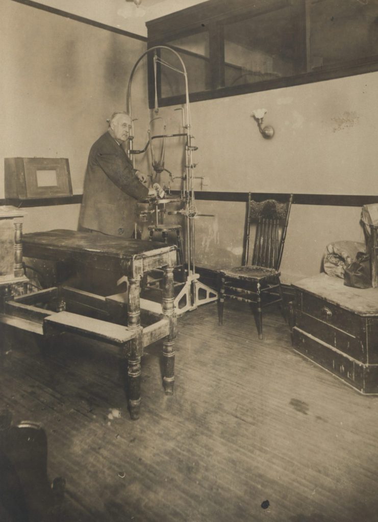 Ridlon stands in a back corner of his office, near some sort of metal apparatus. Also in the room are various pieces of wooden furniture. 