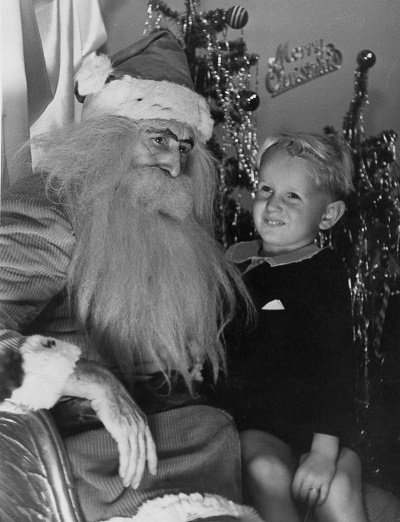 A child in a dark sweater and shorts smiles while sitting on Santa's lap. A Christmas tree with tinsel stands behind them.