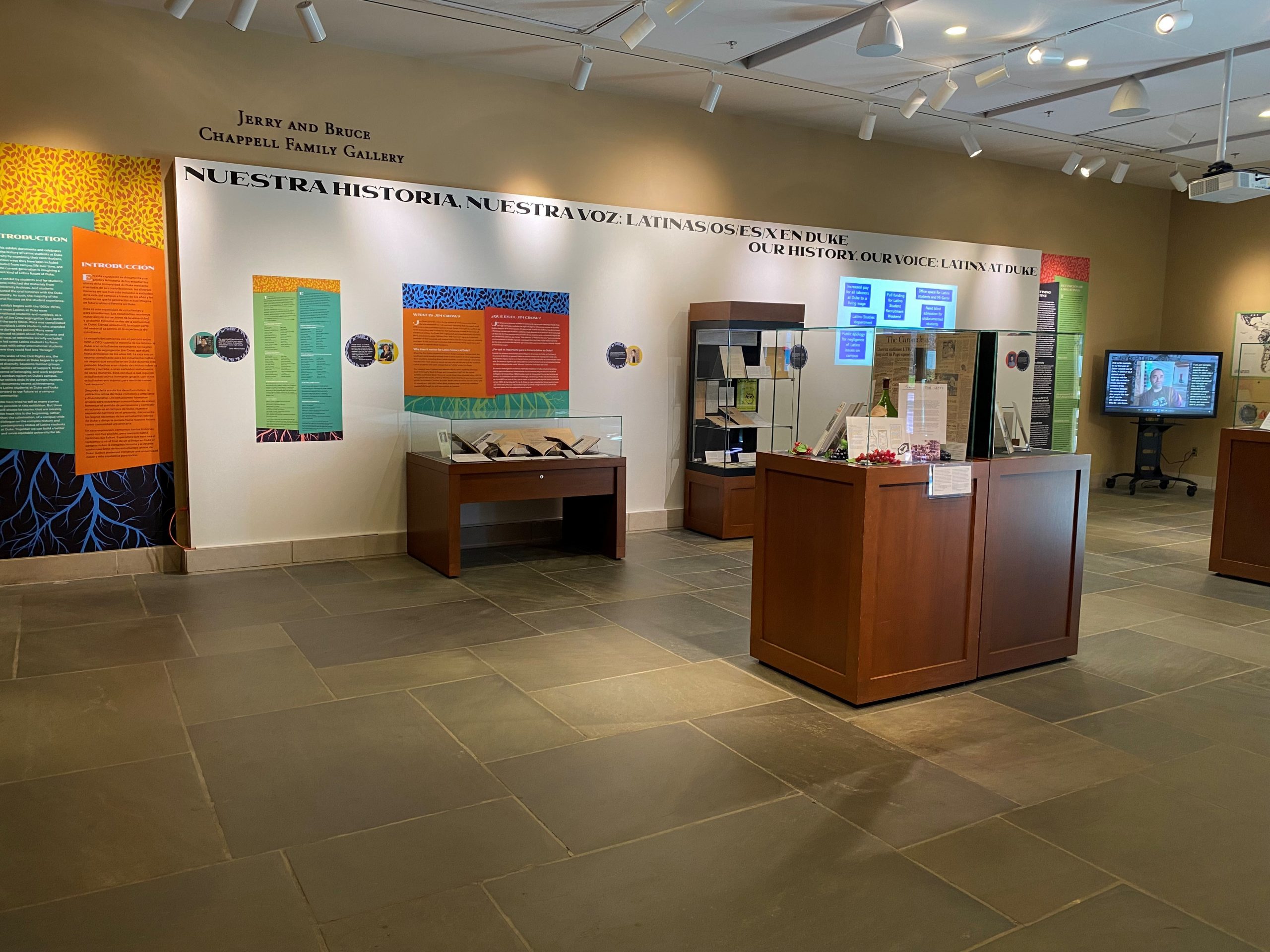 Photograph of the "Our History, Our Voice" exhibit. The exhibit's title appears on the far wall, which is also lined with colorful exhibit panels and exhibit cases. Two exhibit cases display materials in the center of the room.
