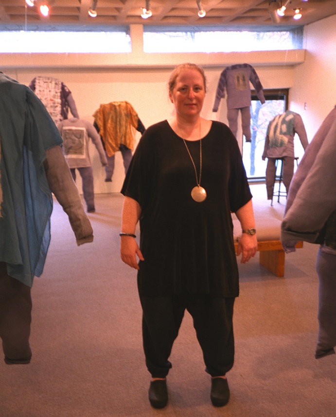 Photograph of Eve Kosofsky Sedgwick. She is wearing a black top and pants, with a gold necklace with a large pendant. She is standing in a gallery that features fabric sculptures in the shape of bodies without heads, arms, or feet. 