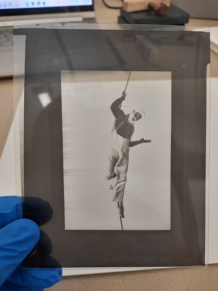 A blue-gloved hand holds a vertically-oriented 4 x 5" negative in a plastic sleeve. The negative shows performer Avner the Eccentric climbing a rope. He is wearing a costume of high-waisted pants with suspenders and a bowler hat.