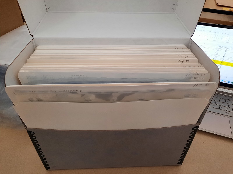 An open gray Hollinger box shows around 30 white paperboard folders, each containing photographic material sleeved in clear plastic.