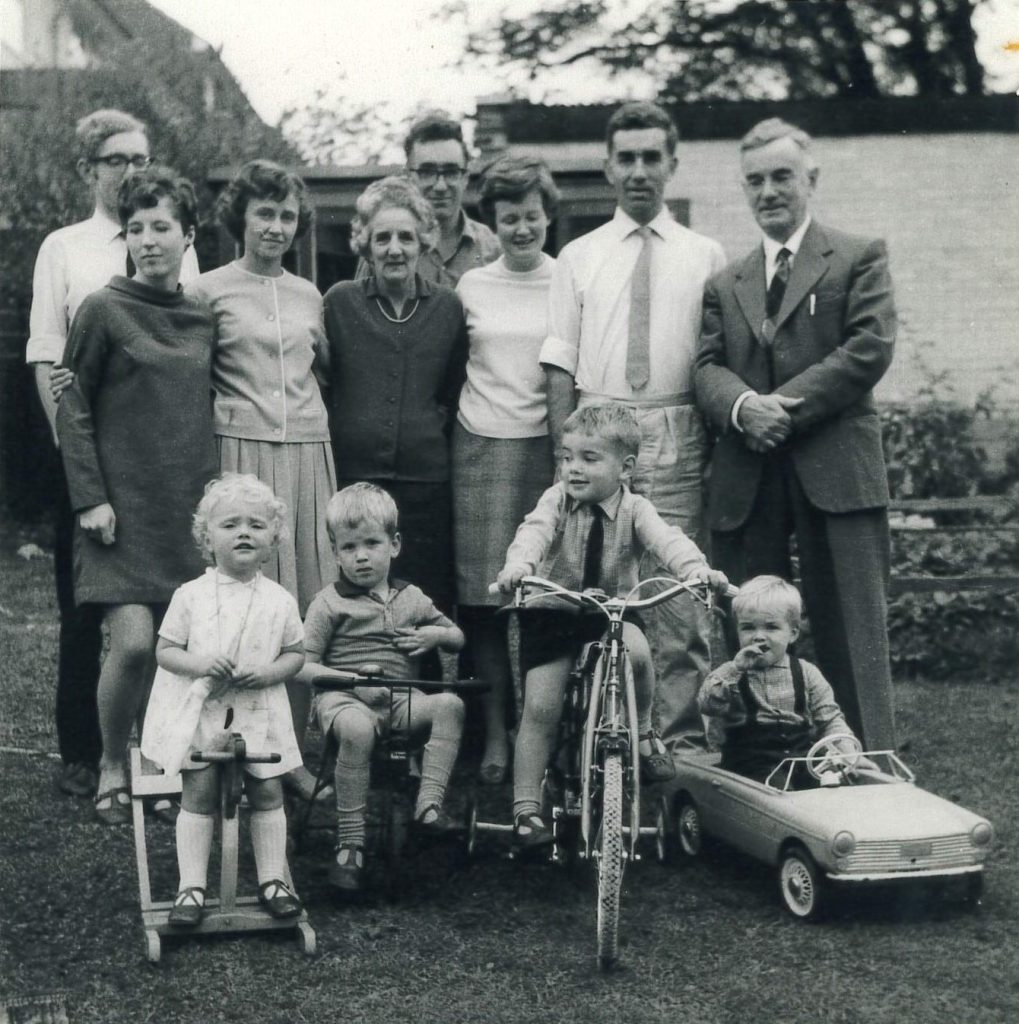 12 members of the Atkinson family pose for a family portrait outdoors, their attire suggesting this might be after a semi-formal event. Four young children are in front on a mix of toy cars, tricycles, and rocking horses.