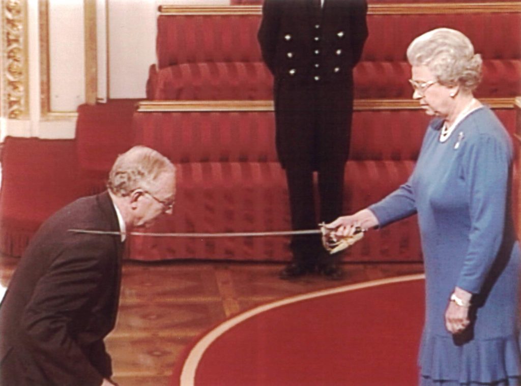 Atkinson kneels before Queen Elizabeth II, who is tapping a saber on his right shoulder.