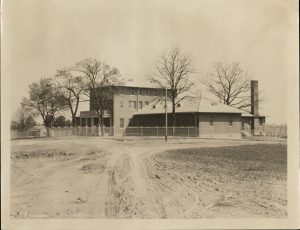 Black and white photograph of a a three story brick building with a single-story wing. The building has a metal fence around it and is in a muddy field. 