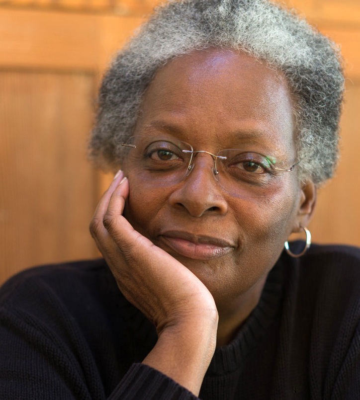 Portrait of Mandy Carter. She is a Black woman, and is seated facing the camera with her chin resting on her hand. She is wearing a black long sleeve shirt and glasses.