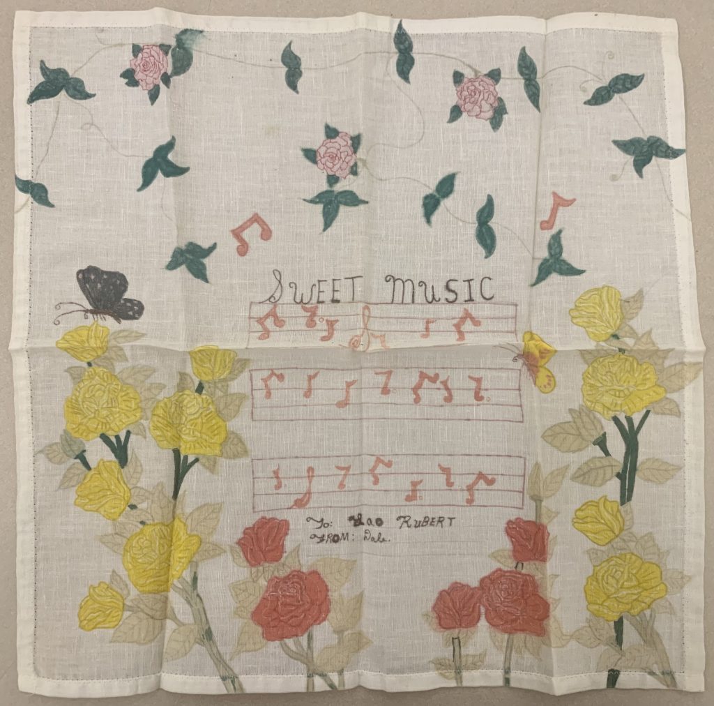 White handkerchief that has been hand-decorated with flowers and butterflies. In the middle there is musical notation with the words "Sweet Music" written above