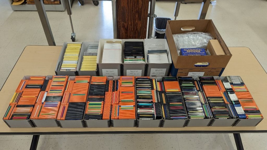 Archival boxes displayed on a table, containing floppy disks, optical disks, hard drives, audio cassettes, and micro cassettes.