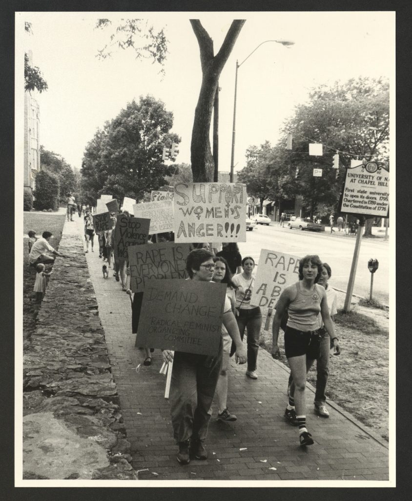 Black and white photograph of a group of women marching a sidewalk on UNC's campus. They are carrying homemade signs, some of the signs read "Support Women's Anger" and "Rape is Everyone's Problem"