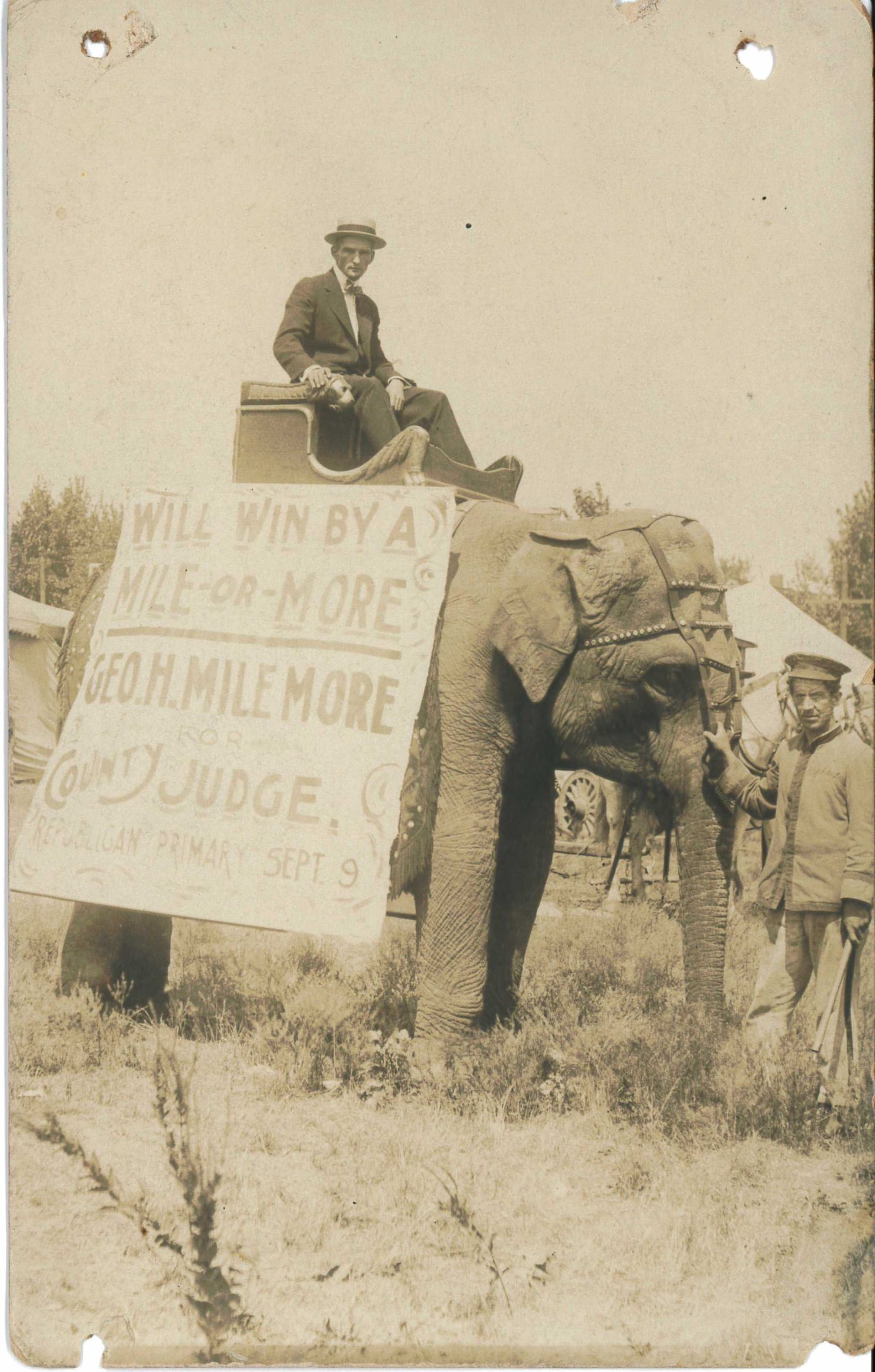 Sepia-toned photograph of an elephant in a grassy field wearing a sign reading "Will Win by a Mile or More. Geo. H. Milemore, County Judge." A man in a dark suit, bow tie, and hat rides the elephant.