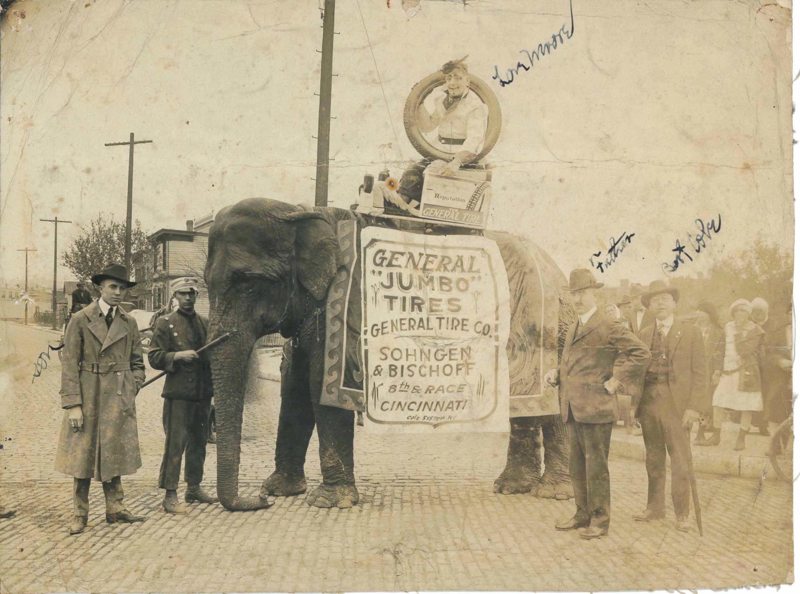 Black and white photograph featuring an elephant wearing a sign reading "General Jumbo Tires, General Tire, Co." A clown, holding a car tire, rides the elephant. The elephant is posed on a paved street with houses in the background and several men in suits standing to the side. 
