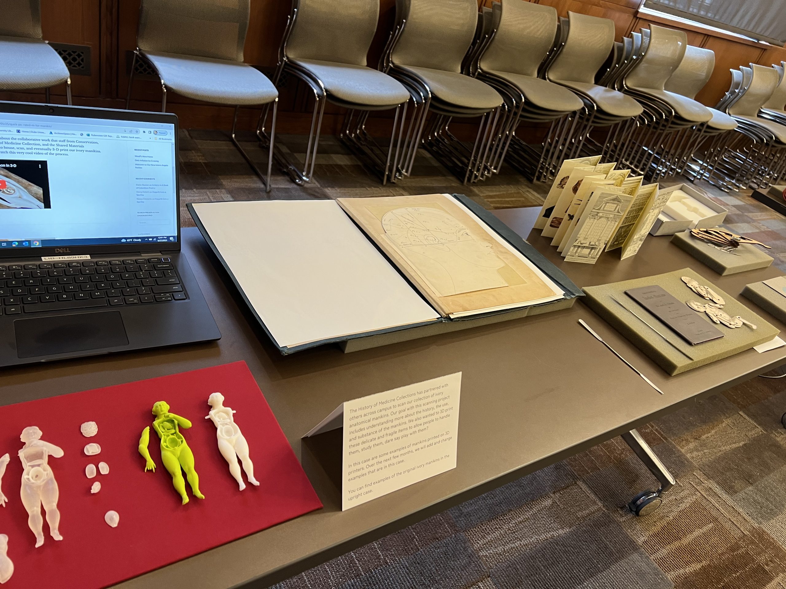 Photograph of table displaying several small, plastic anatomical figures as well as a laptop playing a video showing the making of those figures. The table also displays several books featuring pop-up components. 