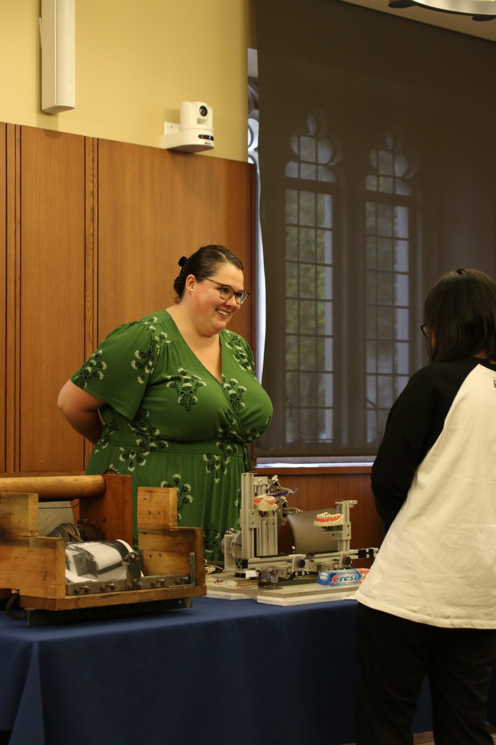 A woman with brown hair wearing a green dress talk with a student, whose back is toward the camera. On a table between the two people, is a toothpaste testing device composed of metal parts and sets of fake teeth. 