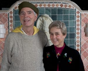 An older man and woman standing in front of an exterior, tiled wall with a wall-mounted fountain.