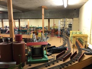 A detailed model train setup in Treynor's two-car garage. The scene features multiple tracks that curve and intersect, with a variety of structures and buildings placed on the layout. There are model trains, including a locomotive, positioned on the tracks. The setup includes industrial elements such as tanks and towers, and there is a bridge spanning some of the tracks. Wooden posts support the ceiling, and the overall layout is intricate and extensive, showing one of Treynor's hobbies. The lighting is provided by overhead fluorescent lights.