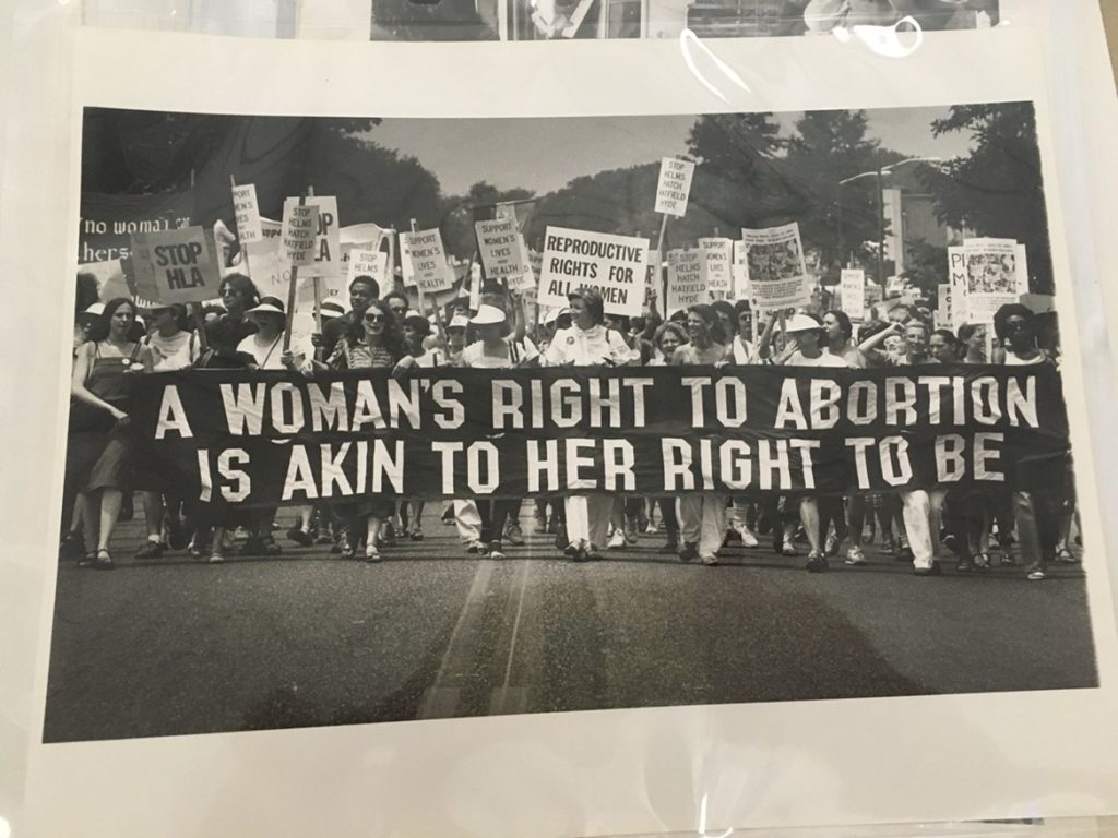 Image of a group of women marching, holding signs and standing behind a banner that reads "A woman's right to abortion is akin to her right to be."