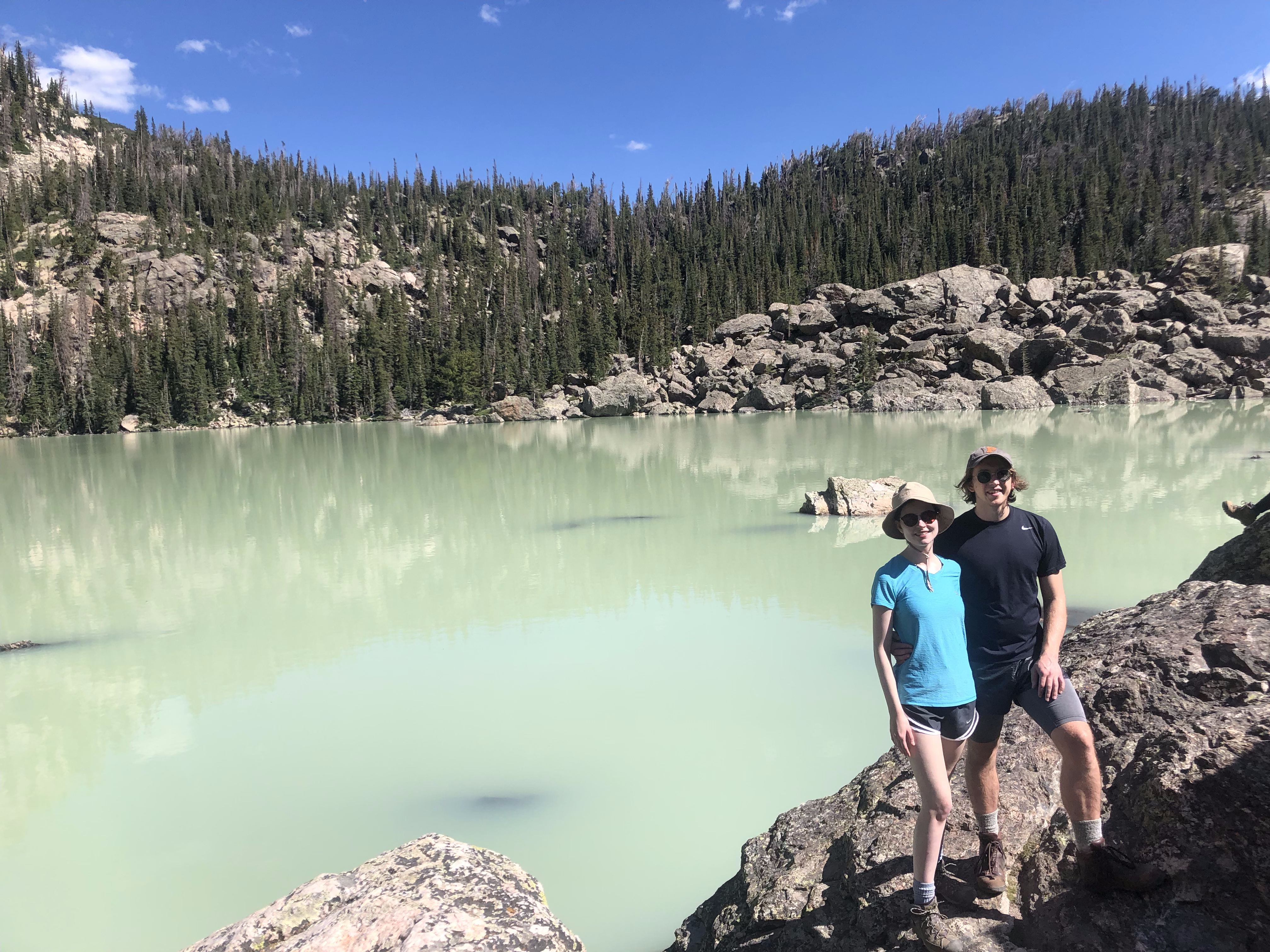 A man and woman stand in front of a pale green lake surrounded by rocks and trees.