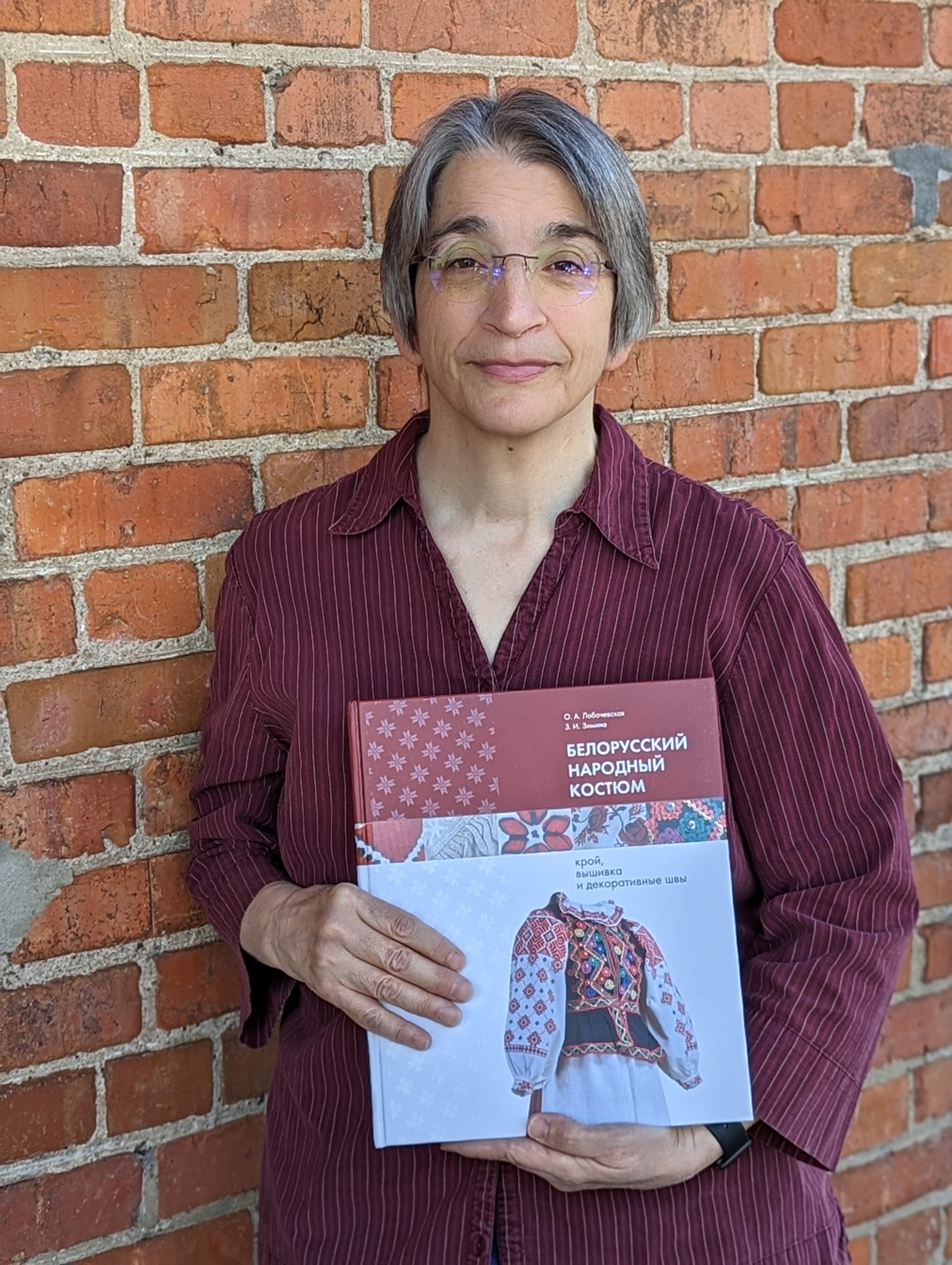 White woman with gray hair in a maroon shirt, holding a book about folk dresses.