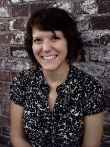 Photo of the blog post author: short brown hair and a big smile, leaning against a white and brown brick wall