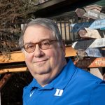 Man in Duke shirt wearing glasses in front of signpost art at home.