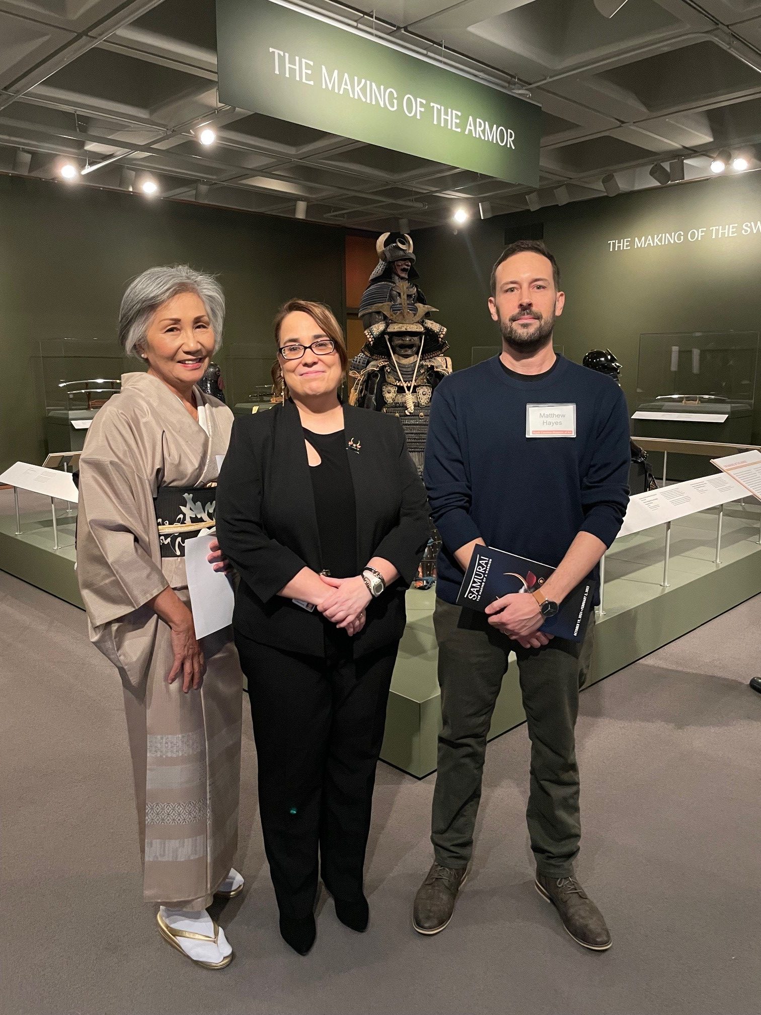 Two women and a man standing in front of samurai armor at a museum exhibition.